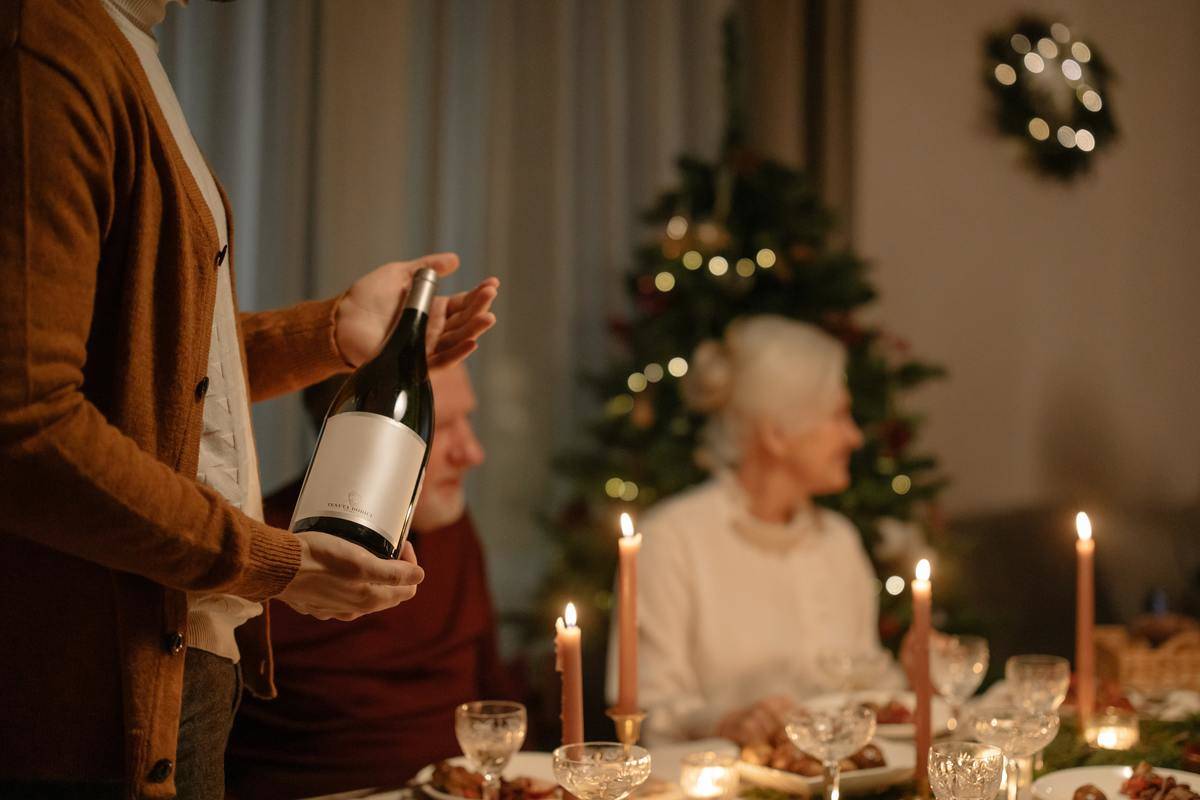 person-standing-while-holding-wine-bottle in front of elderly couple at christmas table