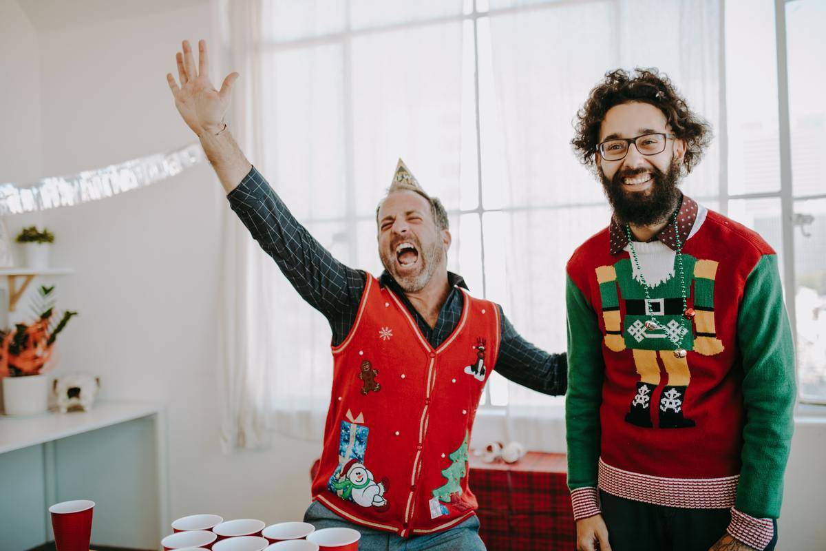Two men in an office setting in funny Christmas sweaters, one cheering with his arm up and one laughing.