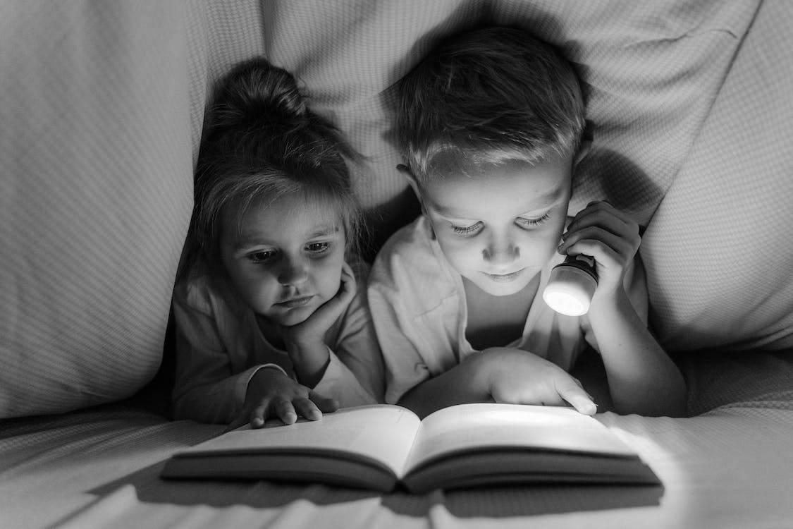 A young boy and girl are reading under a blanket, the boy holding up a flashlight.