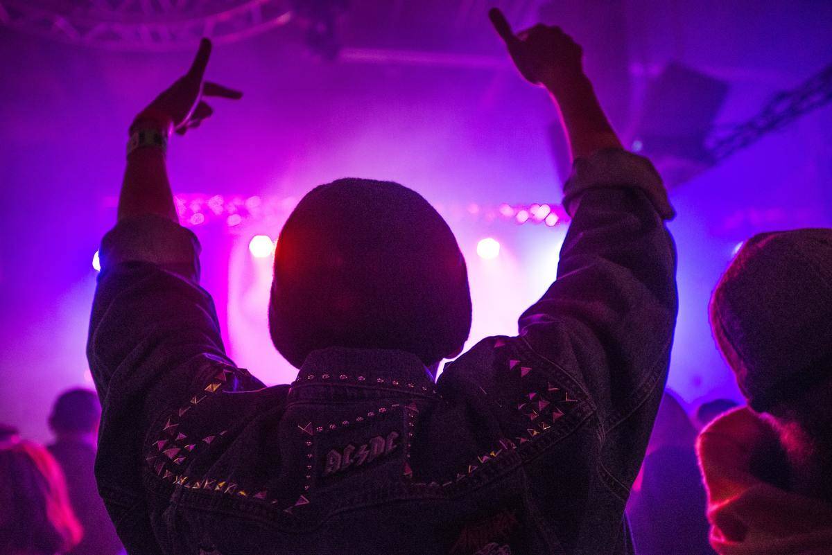 Someone at a concert as seen from behind, hands up with a denim jacket and beanie on, pink lights on stage.