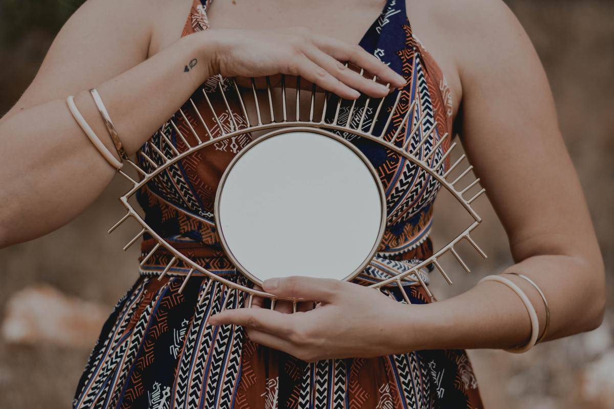 A woman in a pattered dress holding an eye-shaped mirror in front of her chest.