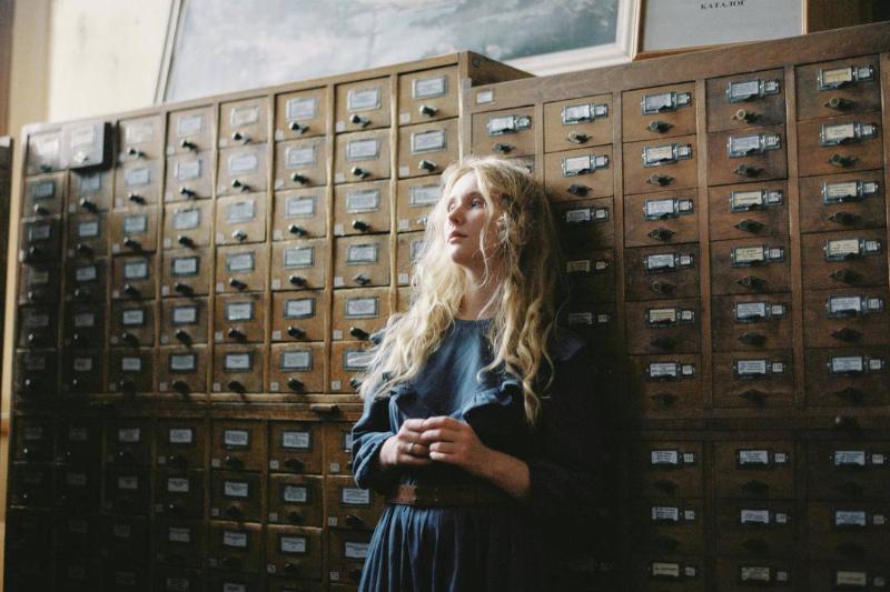 A blonde woman against a cabinet with many small cubbies, looking away whistfully.