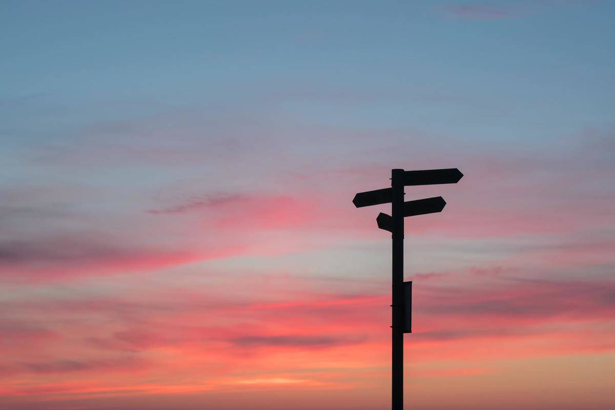 A silhouette of a signpost pointing in four directions against a sunset sky.