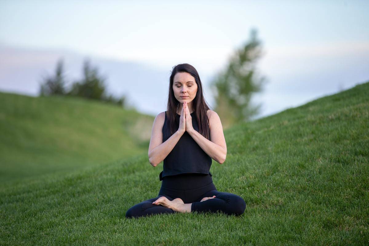 A woman sitting among grassy hills, legs folded and hands together, meditating.