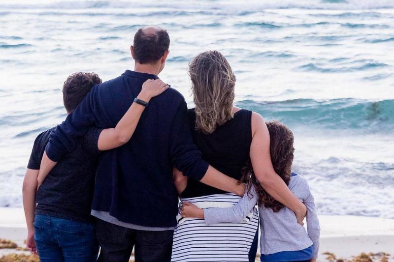 A family standing on a beach, facing the water, all with their arms around each other.