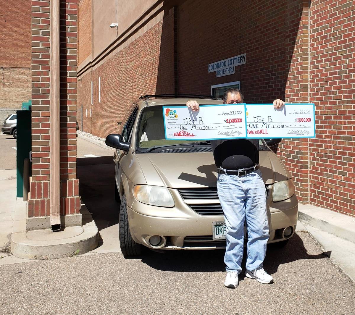 Joe standing in front of his car, holding up his two $1 million checks.