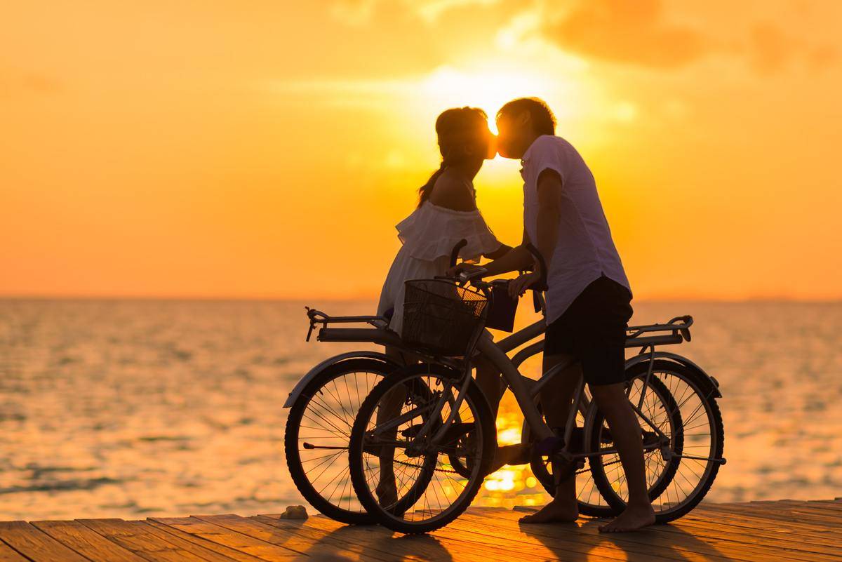 A couple each atop a bike, kissing on a boardwalk in front of a sunset above the water.
