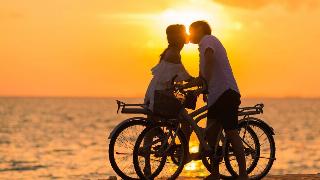 A couple each atop a bike, kissing on a boardwalk in front of a sunset above the water.