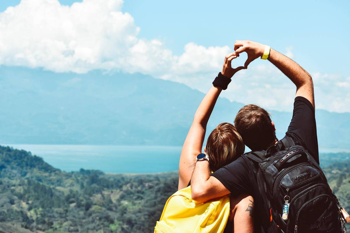 A couple standing atop a mountain, facing away from the camera, each using one hand to make a heart shape above their heads.