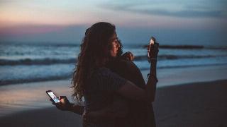 couple-hugging-and-using-smartphone-near-sea-on-sunset