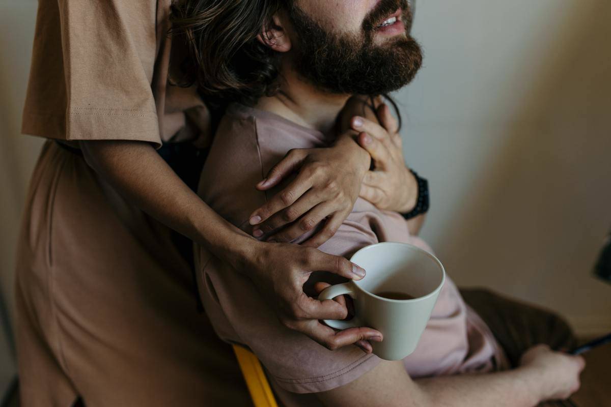A man is sitting in a chair while a woman embraces him from behind while standing and holding a mug.