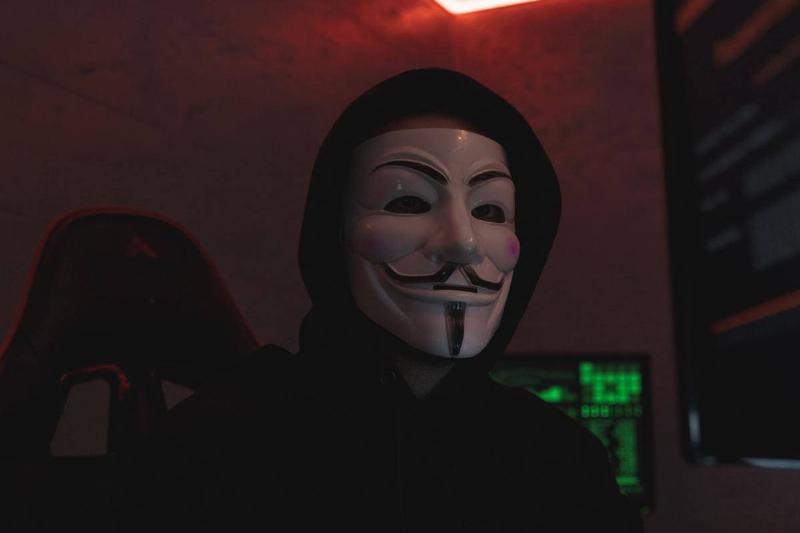 person in hoodie and anonymous mask sits at computer with code