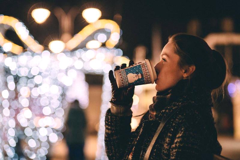 A woman drinking from a coffee mug outside in front of some Christmas lights.