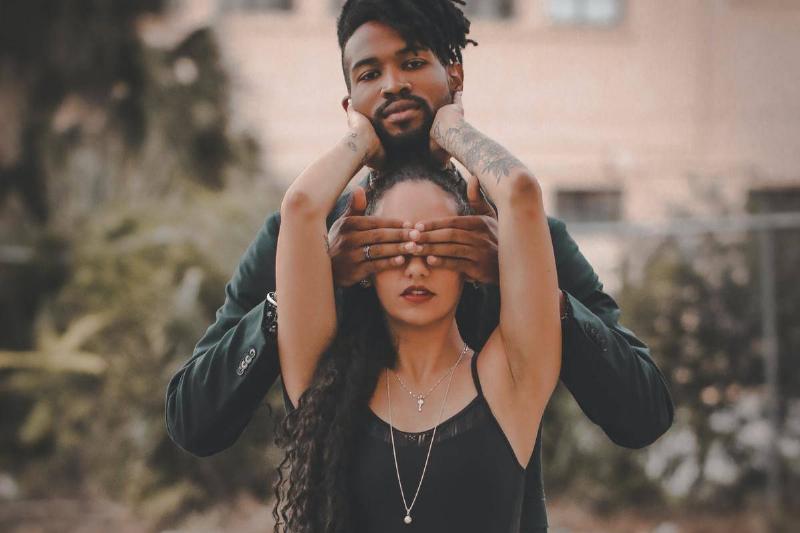 woman stands in front of man holding his face while he covers her eyes