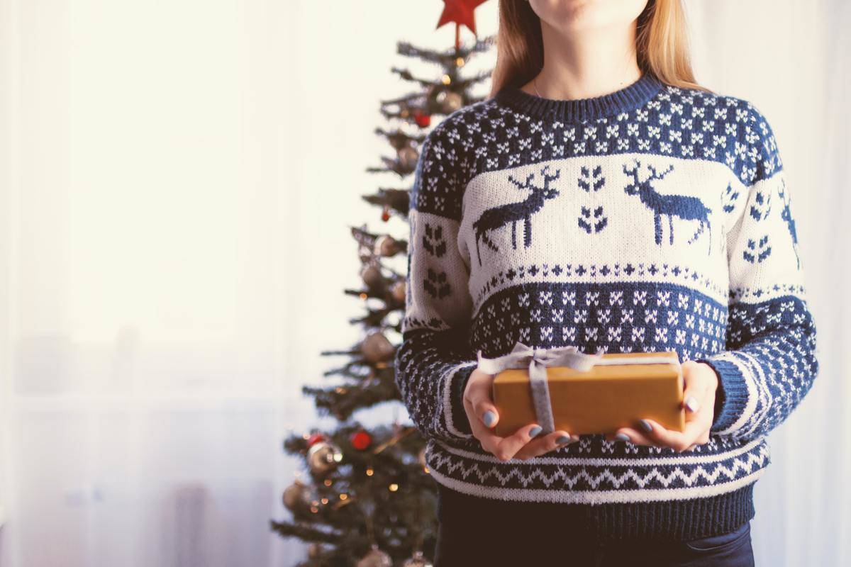 A woman in a blue and white Christmas sweater holding a small present while standing in front of a Christmas tree.