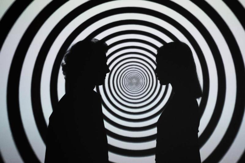 Silhouettes of two people facing one another in front of a black and white spiral background.
