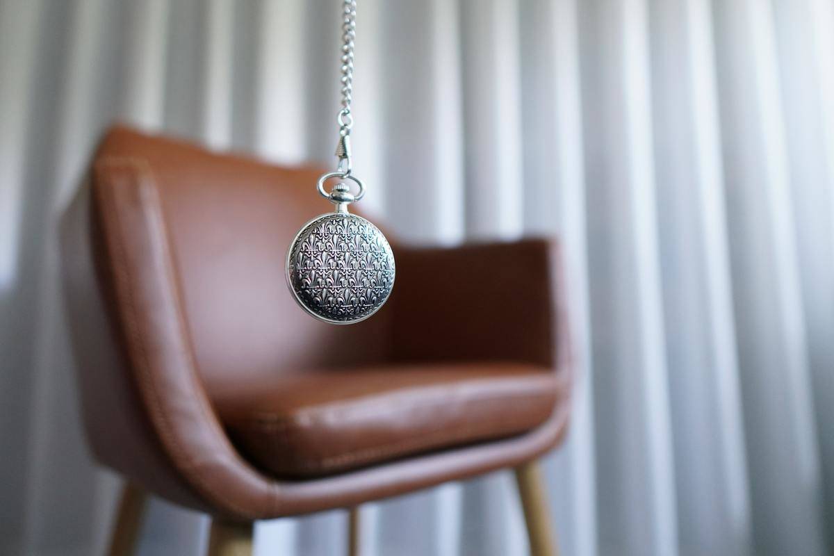A silver pocketwatch in focus in the foreground, with a brown leather chair out of focus in the backgorund.