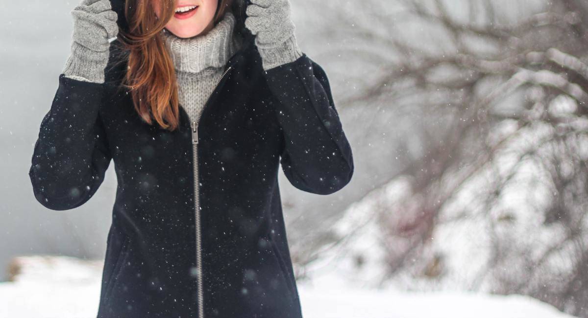 A woman walking in the snow, pulling her coat hood up.