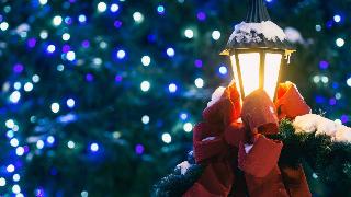 A streetlamp decorated with a big red bow and some garland, dusted with snow, in front of some Christmas lights.