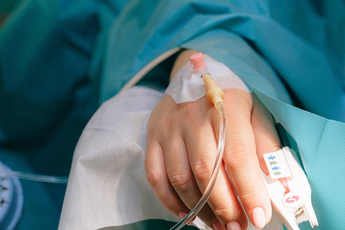 A hand with an IV needle inserted and a heart monitor on their thumb.