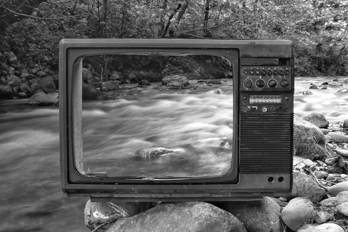 A greyscale image of an old TV frame with no screen propped up in front of a creek.