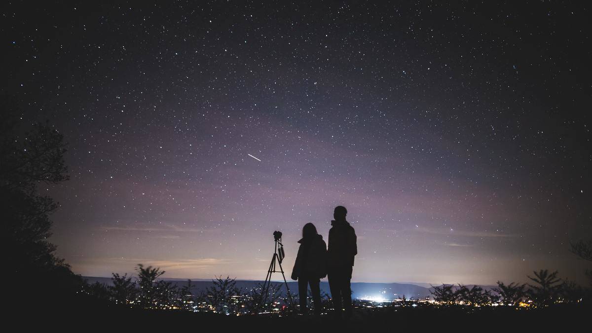 A silhouette of a couple standing next to a telescope before a sky full of stars.