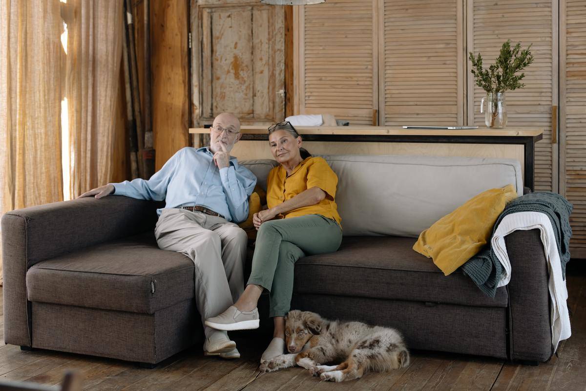 An elderly couple sitting on a couch with their dog at their feet.