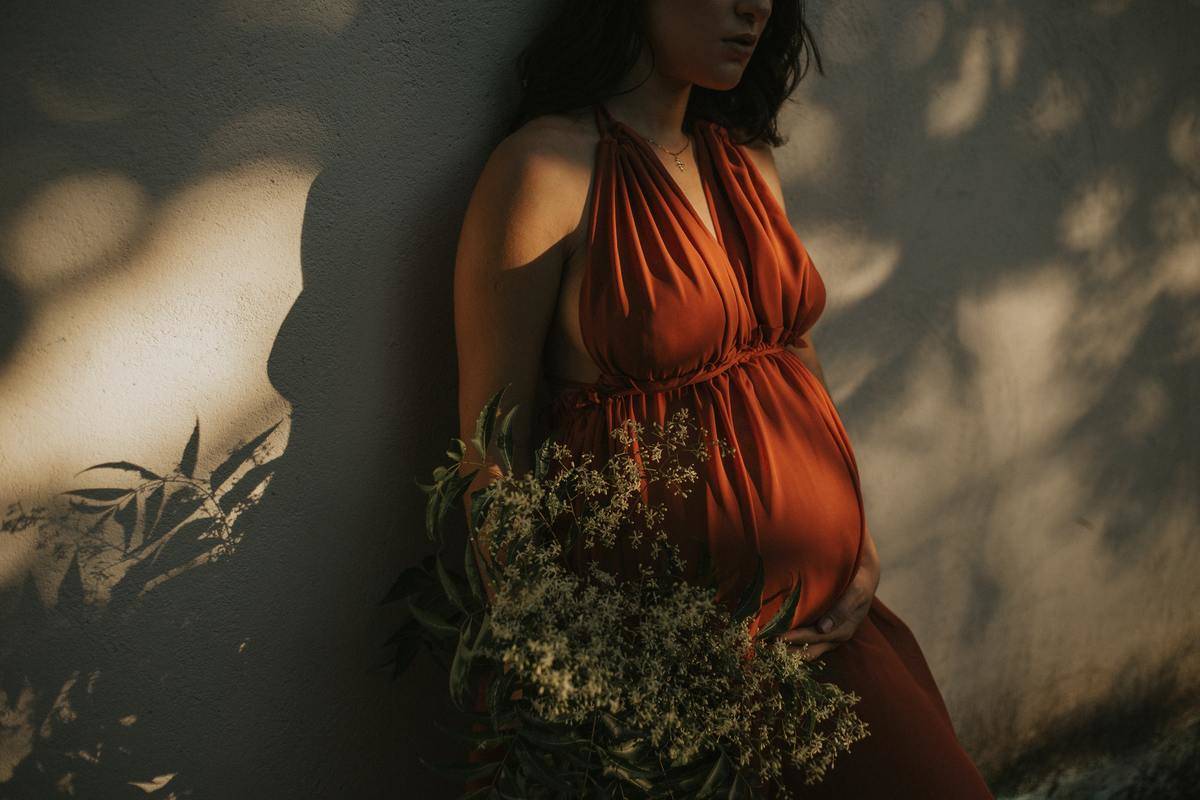 A pregnant woman in an orange dress leaning against a wall, holding a bouquet.