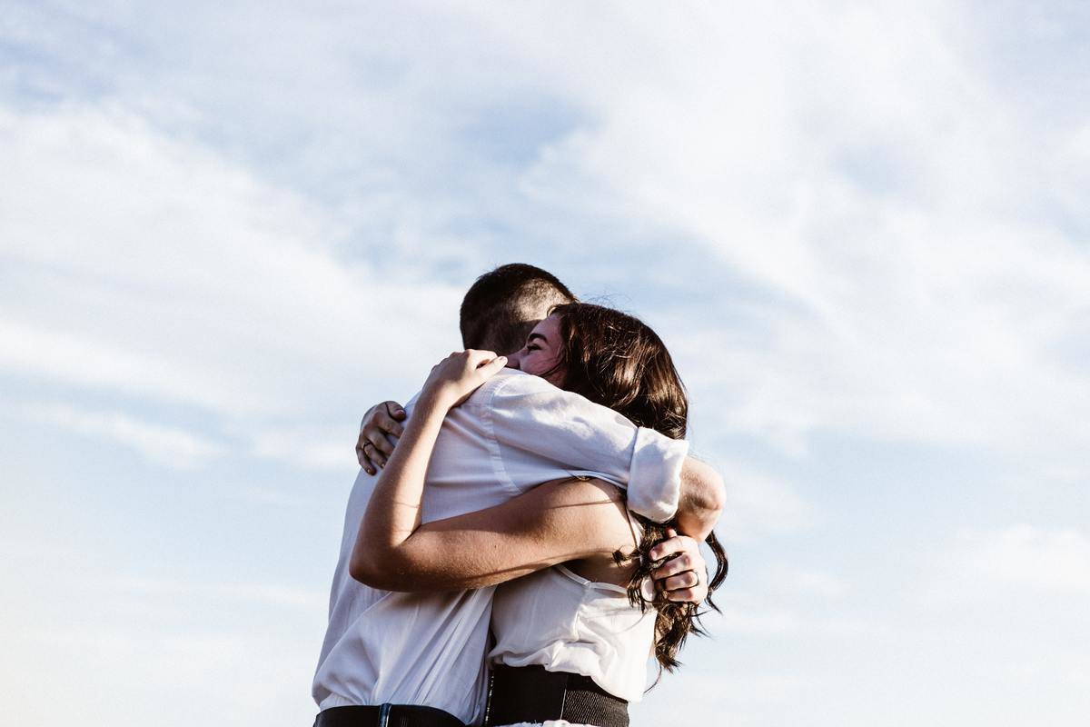 A man and a woman in white hugging in front of a pale blue sky.