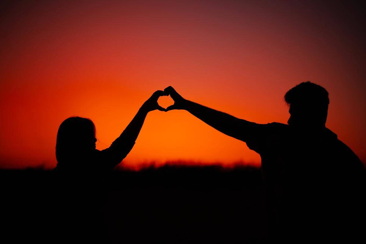 A silhouette of a couple in front of a sunset, the two making a heart shape with their hands.