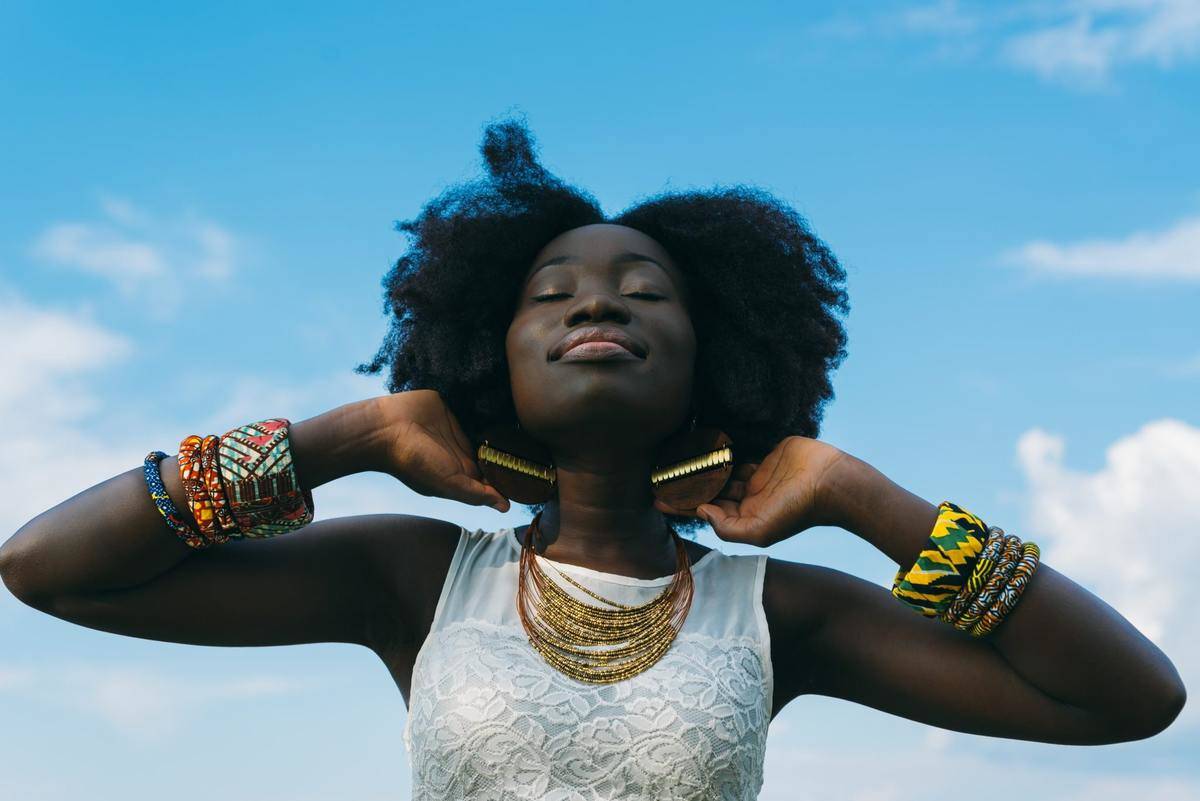 A woman smiling in front of a blue sky background, hands raised to push her large earing forward, smiling with her eyes closed.