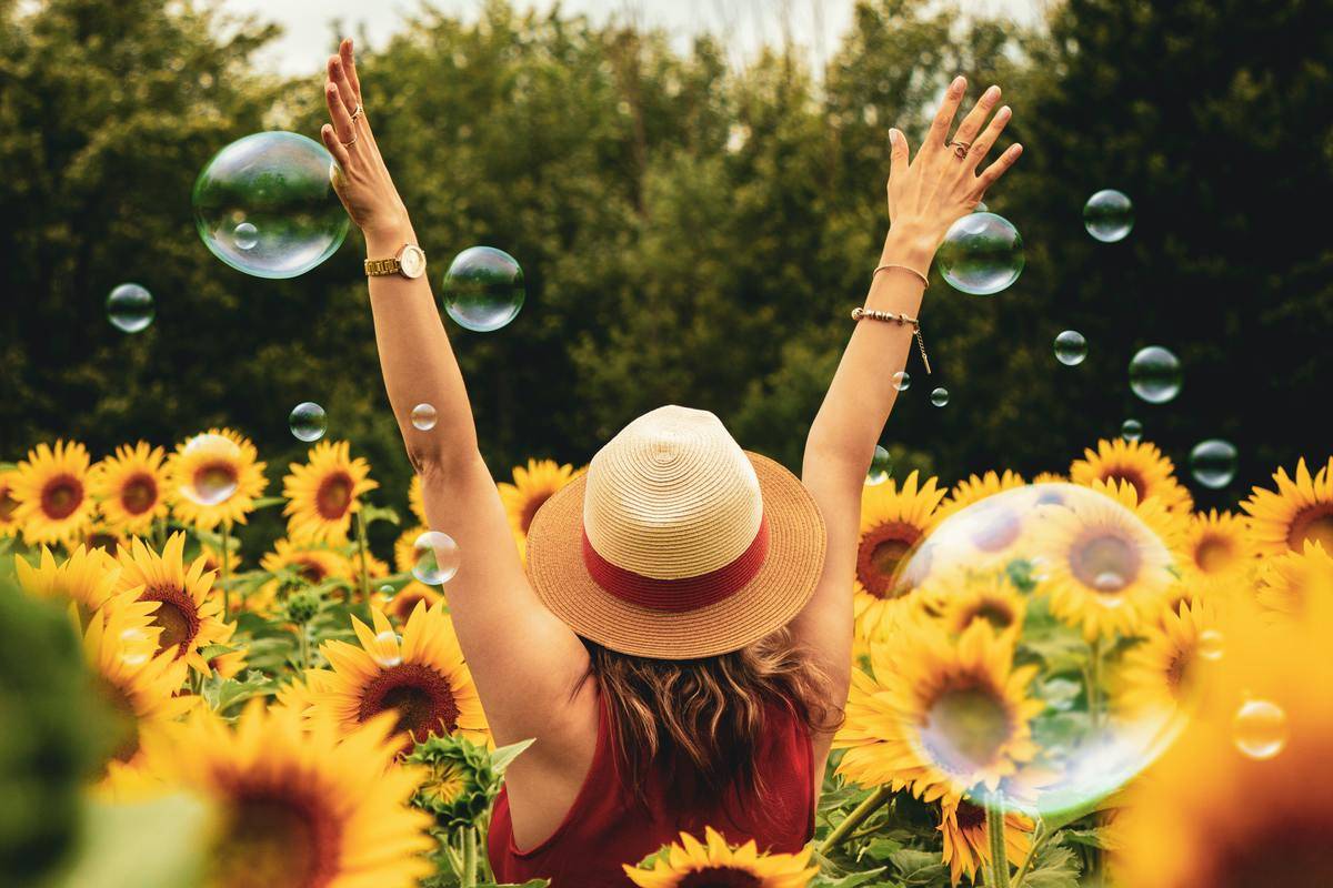 A woman standing with her arms up in a field of sunflowers, bubbles floating around her.