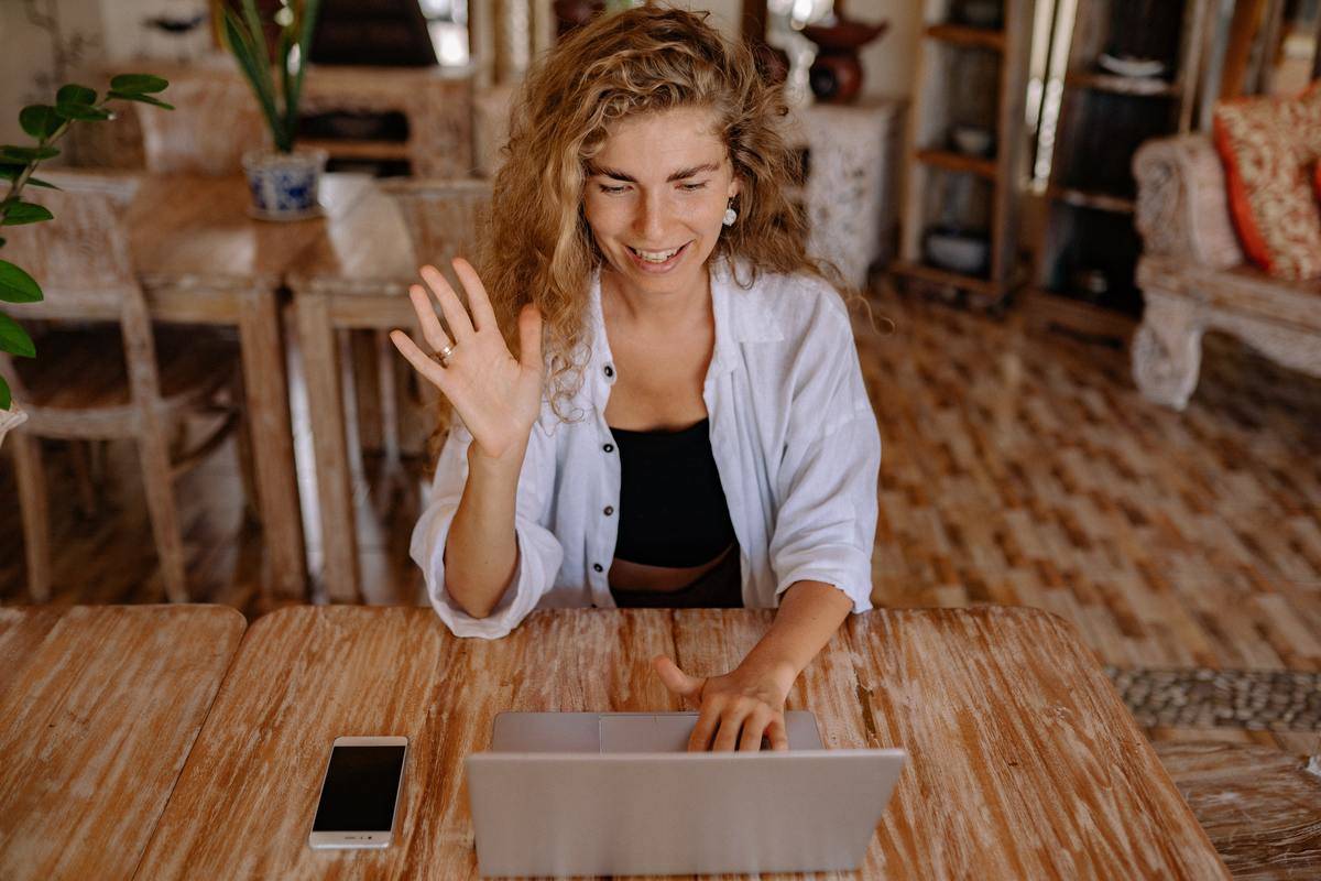 A woman sitting in front of her laptop, raising a hand in greeting toward someone she's on a video call with.