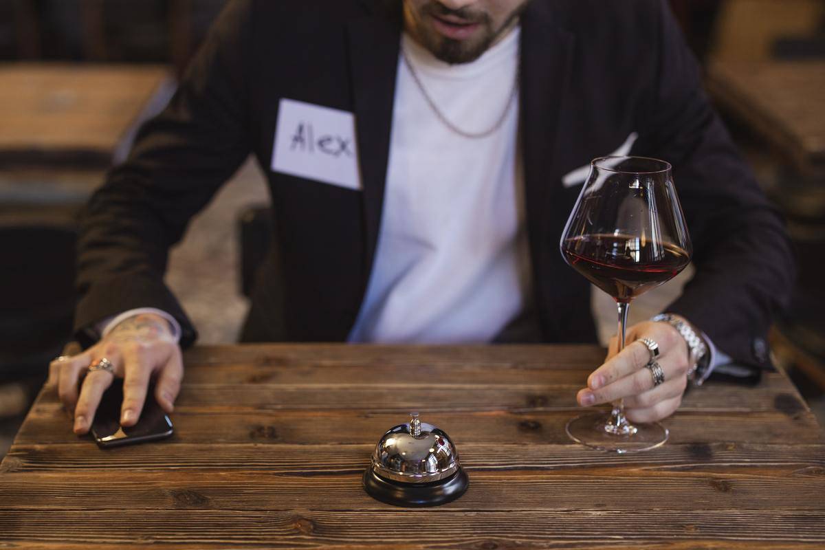 A man sitting at a table at a speed dating event, phone in one hand, wine glass in the other, a bell on the table.