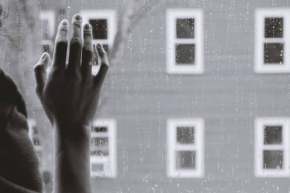 A greyscale image of someone's hand pressed longingly up to a rain-covered window.