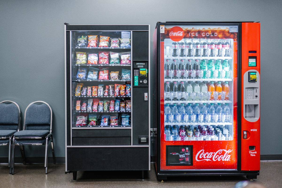 Two vending machines against a grey wall, one for snacks and one coca-cola branded one for drinks.