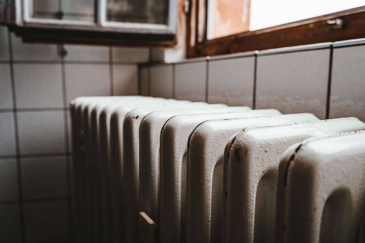 A white radiator against a white tile wall, below a window.