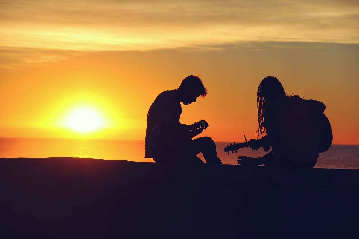A silhouette of two people playing guitar against a sunset sky.