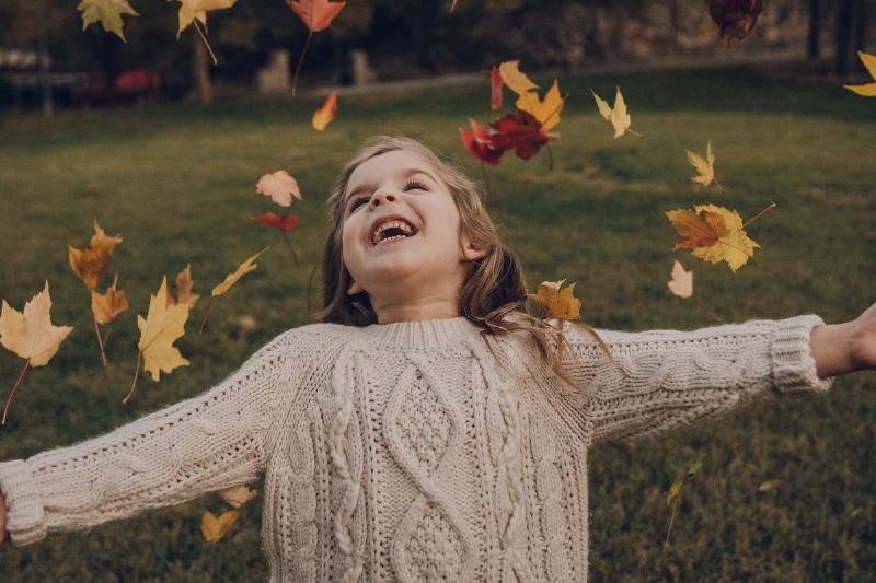 A young girl extending her arms as she looks upwards, smiling, autumn leaves falling around her.