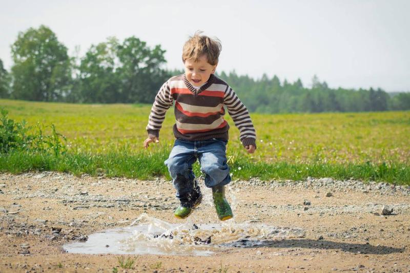 A young boy jumps in a puddle on a dirt road.