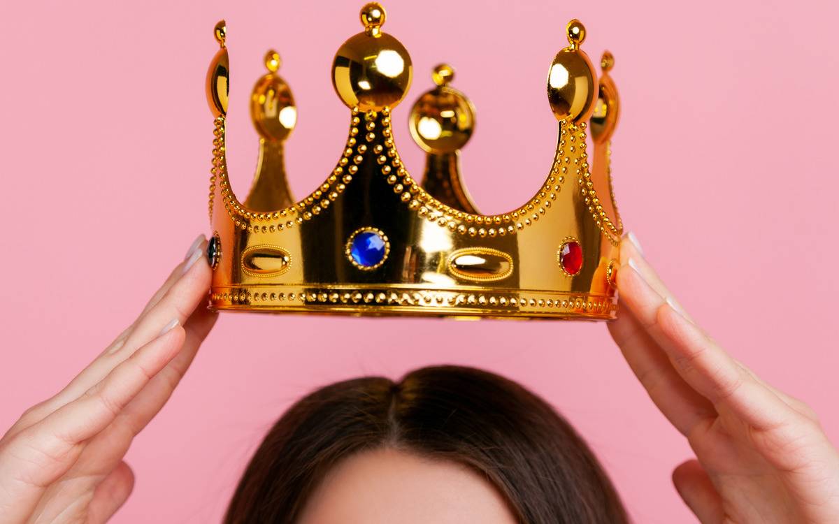 A woman holding a gold crown just above her head against a pink background.