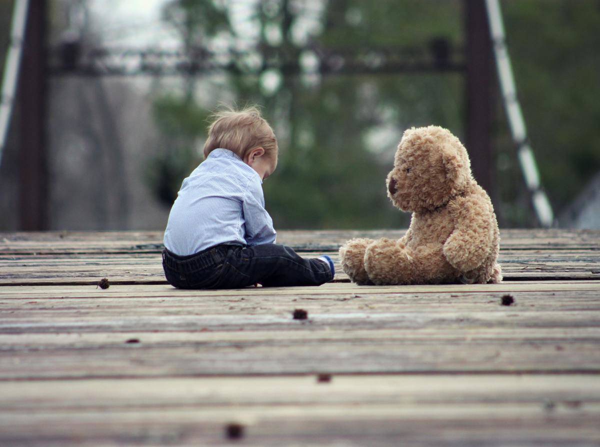 A very young boy sitting on a wooden bridge next to a teddy bear his size.