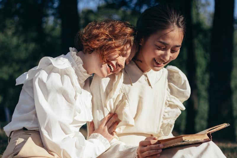 Two women sitting on a blanket outside, smiling, one holding a book and the other leaning against her shoulder.