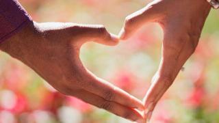 Two hands making a heart shape together against a blurred pink and green backdrop.