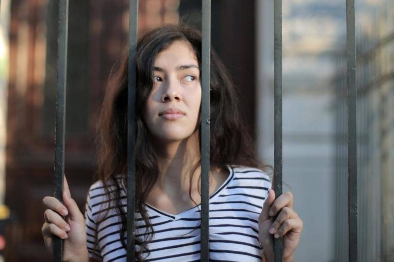 A girl standing behind a barred fence, clutching the bars and looking off to the side.