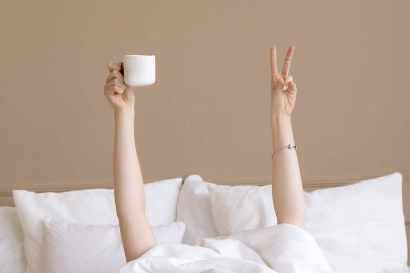 Someone under their blankets, arms raised up from underneath, one holding a coffee cup and the other making a peace sign.