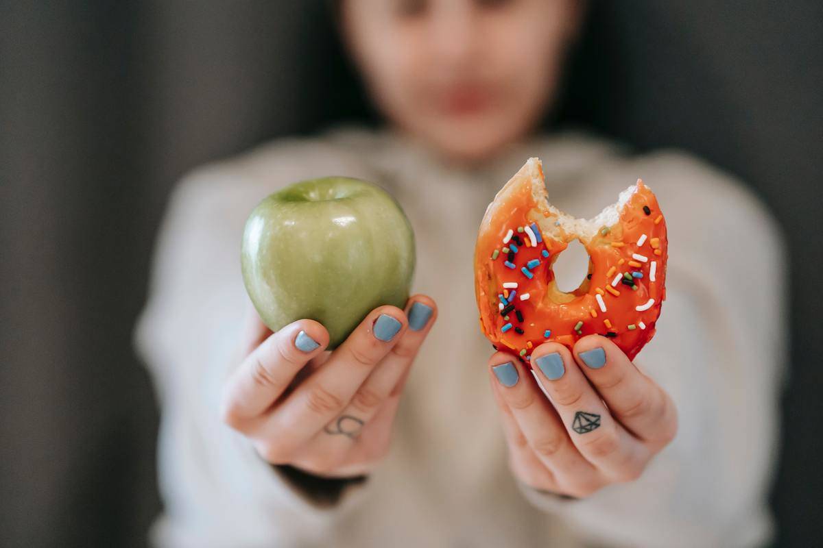 A woman holding up an apple in one hand, and a donut with a bite taken out of it in the other.