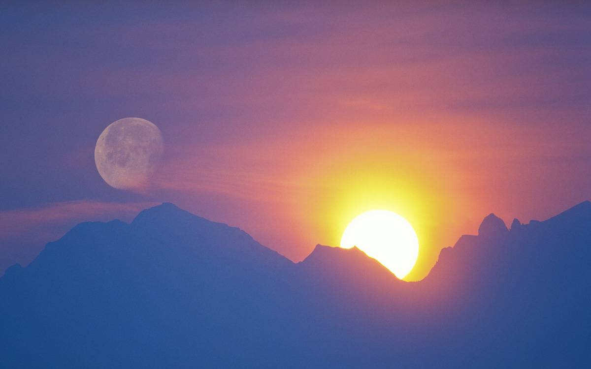 The sun rising over a mountain range with the moon in view to the left.