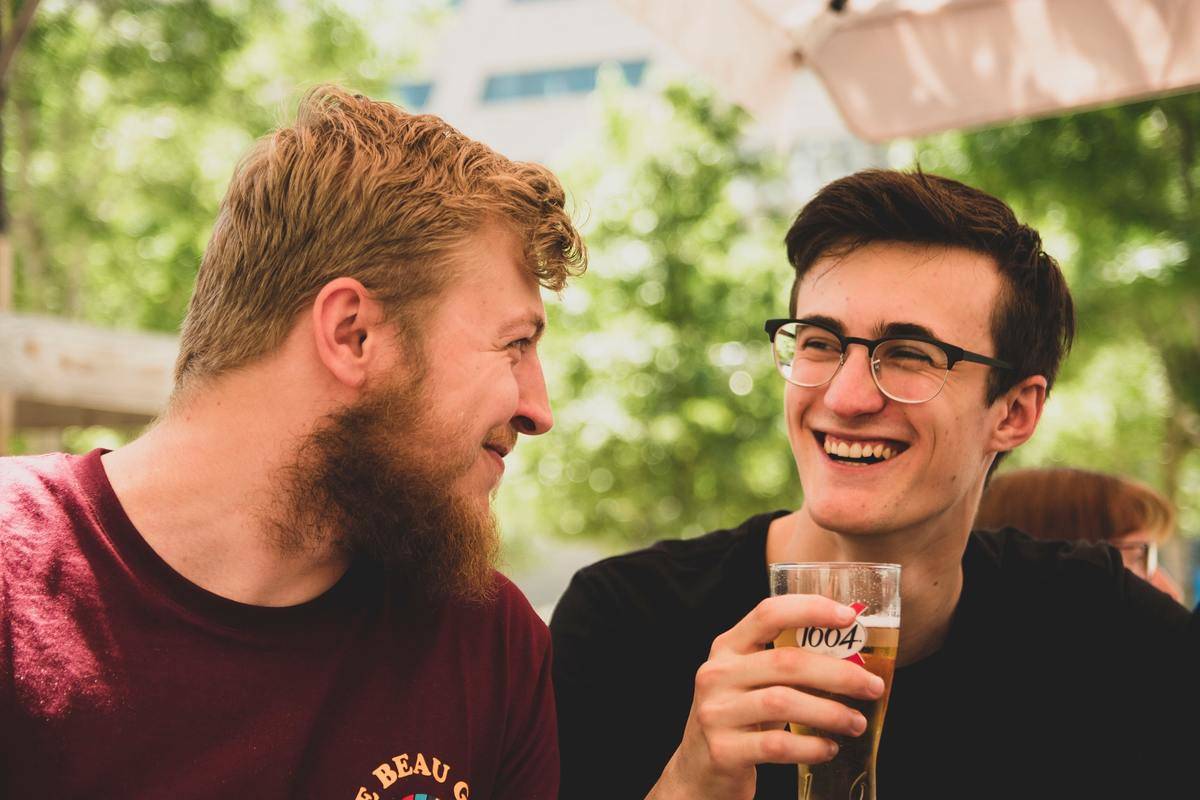 Two friends sitting and smiling at each other, one holding a beer.