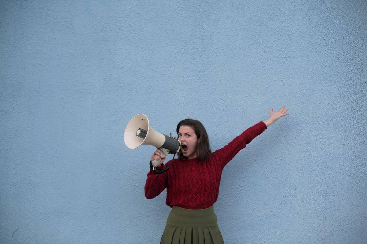 A woman against a blue wall yelling into a megaphone, her other arm outstretched upwards.